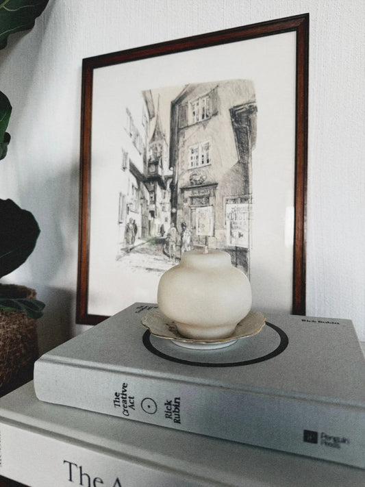 Dekokerze in Form eines japanischen Teegefässes auf einem Stapel von Büchern. Dahinter ist ein Bild zu sehen.