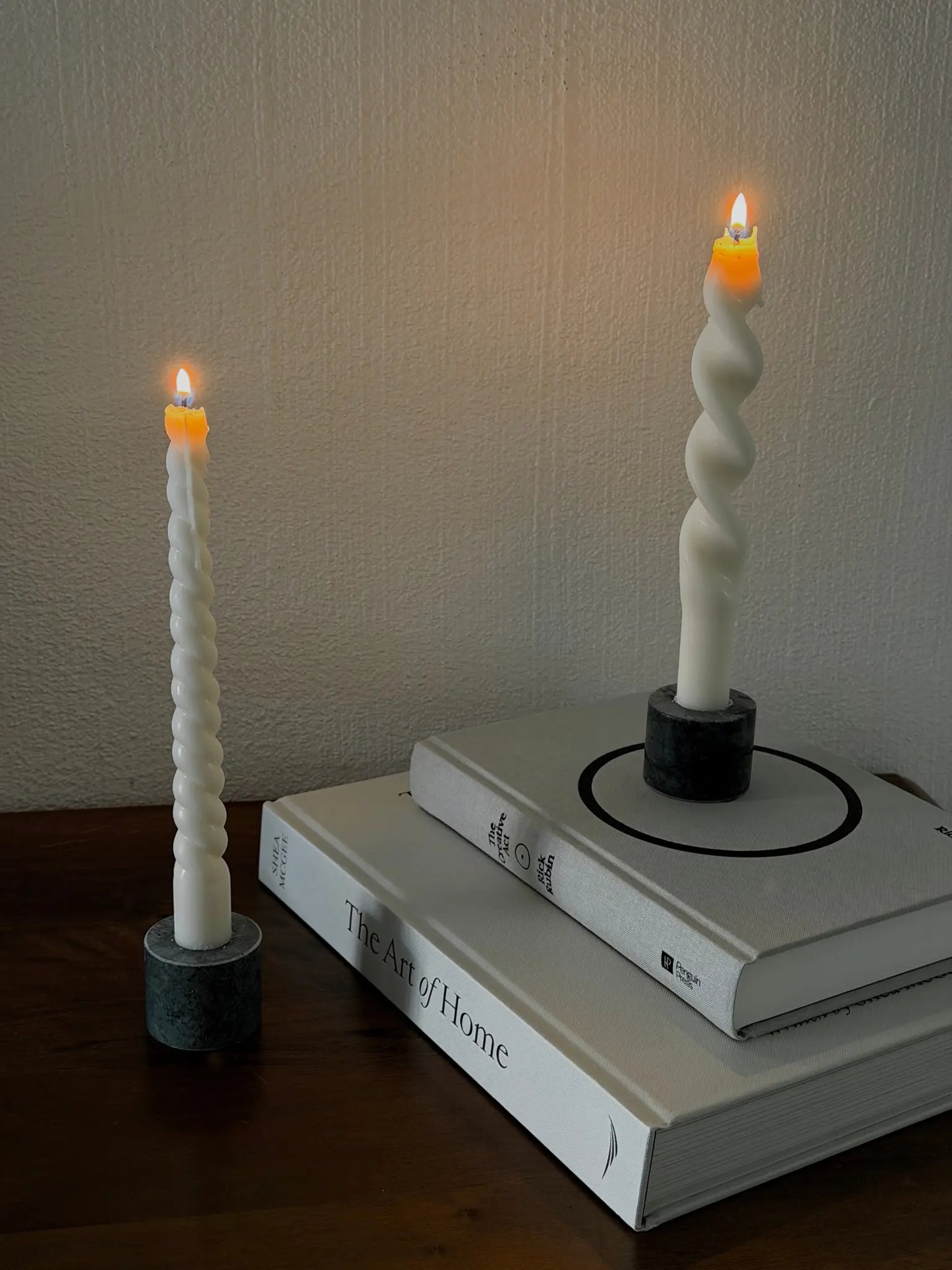 Zwei verschieden gedrehte, brennende Stabkerzen auf einer Holzkommode. Einer Stabkerze steht auf zwei Büchern.