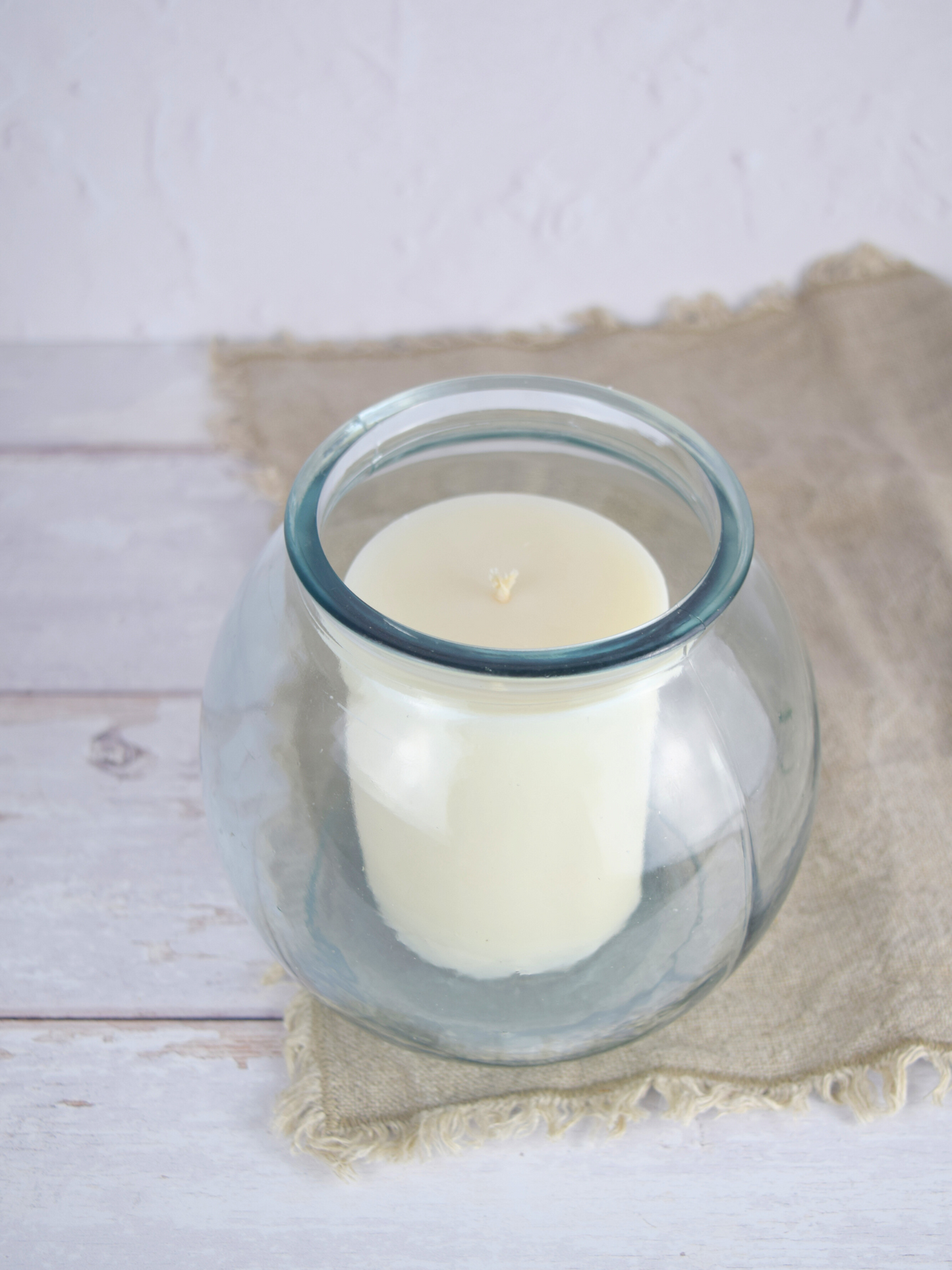 Durchsichtige, runde Vase mit einer weissen Zylinderkerze auf einem braunen Tuch vor einem weissen Hintergrund
