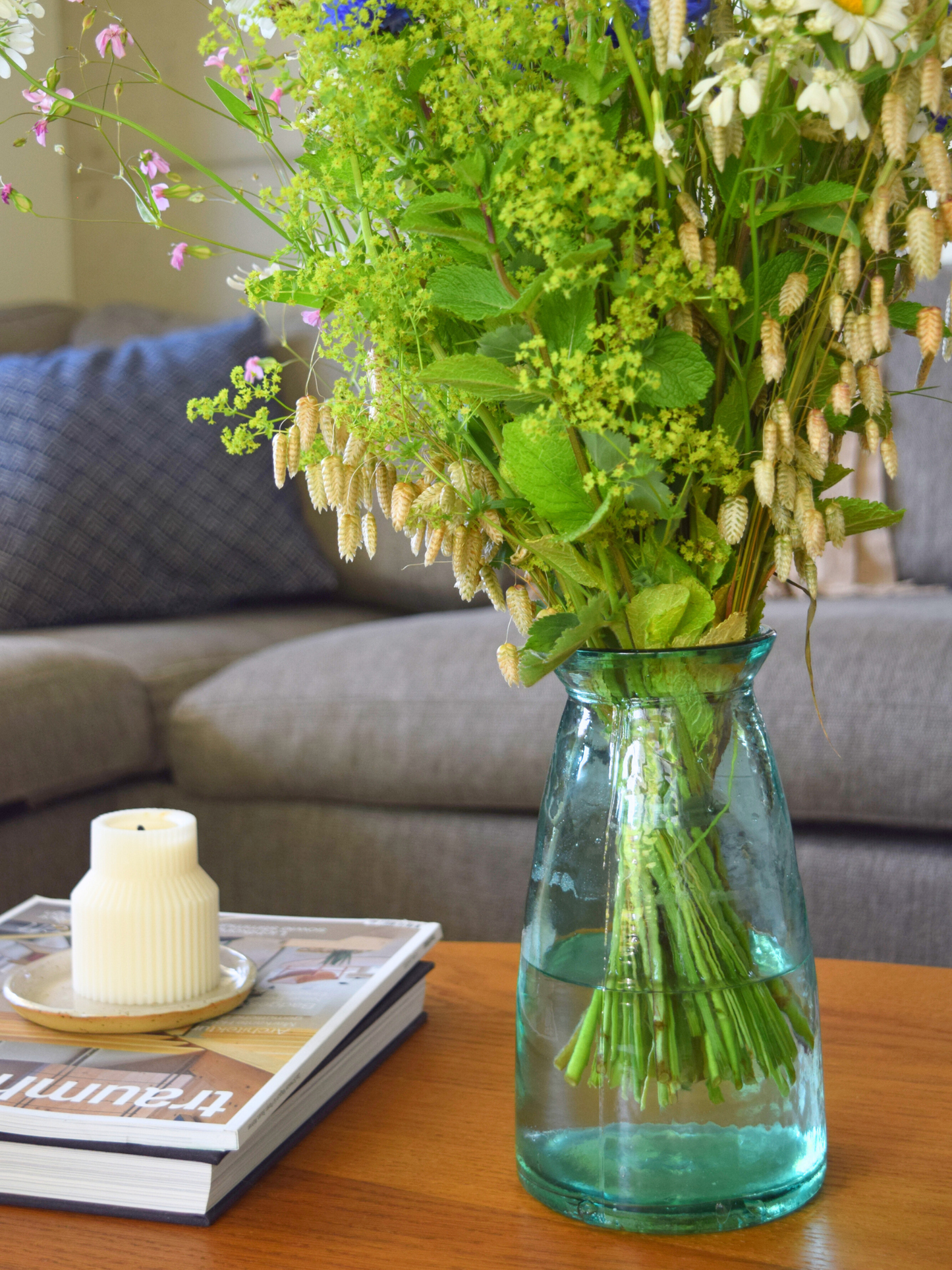 Bläuliche, grosse Vase mit einem Blumenstrauss stehend auf einem Holztisch mit einer Kerze. Im Hintergrund steht ein graues Sofa.