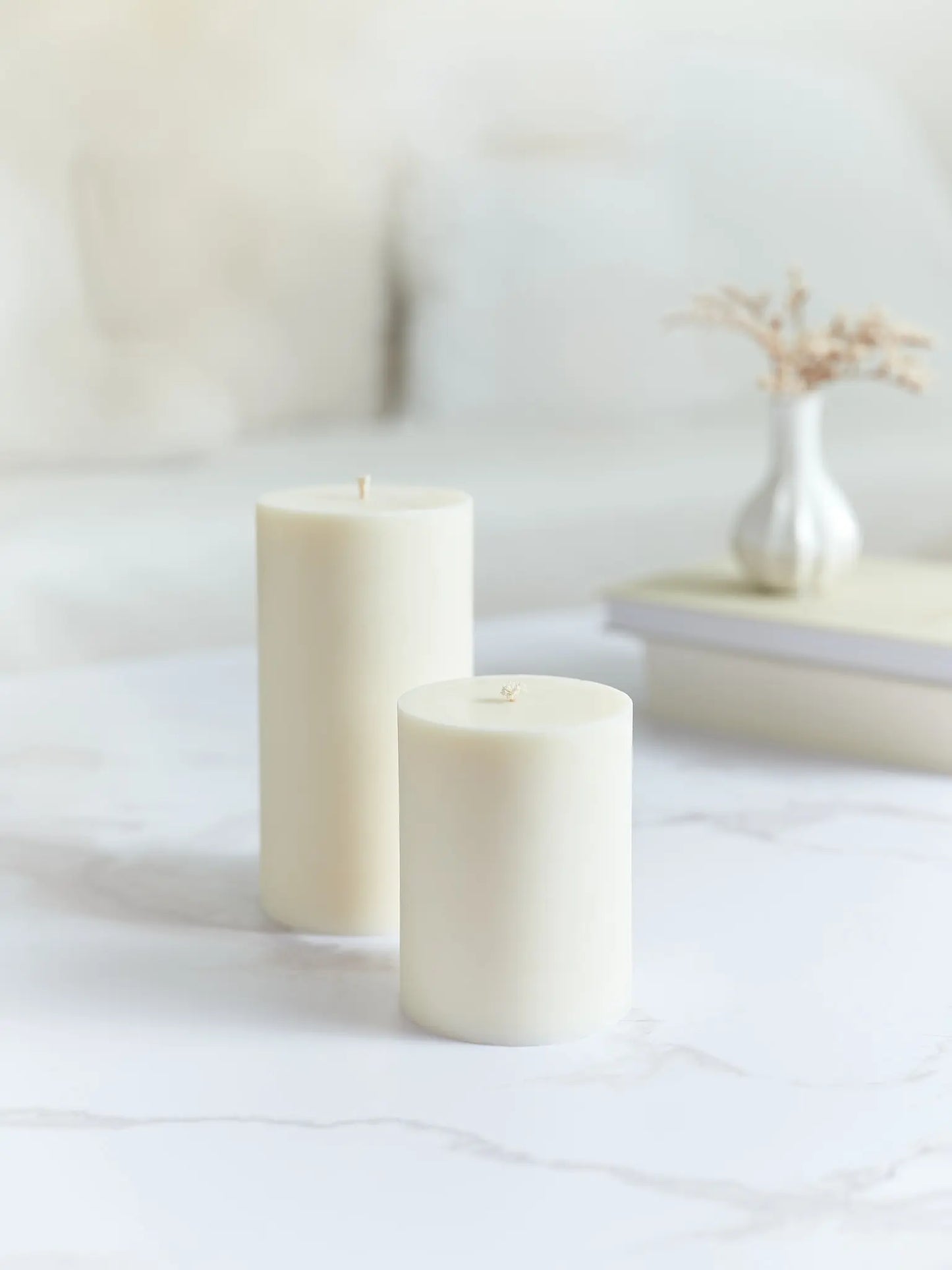 Eine grosse und eine kleine glatte Zylinderkerze in cremeweiss. Beide stehen auf einem Marmorcouchtisch und im Hintergrund hat es Bücher und eine Vase mit Trockenblumen.