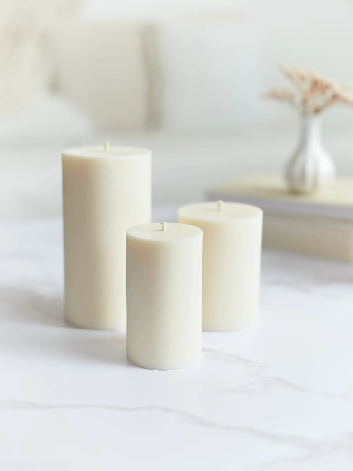Drei cremeweisse, glatte, unterschiedlich grosse Zylinderkerzen auf einem Marmorcoutisch mit einer Vase und Büchern im Hintergrund.