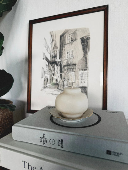 Dekokerze in Form eines japanischen Teegefässes auf einem Stapel von Büchern. Dahinter ist ein Bild zu sehen.