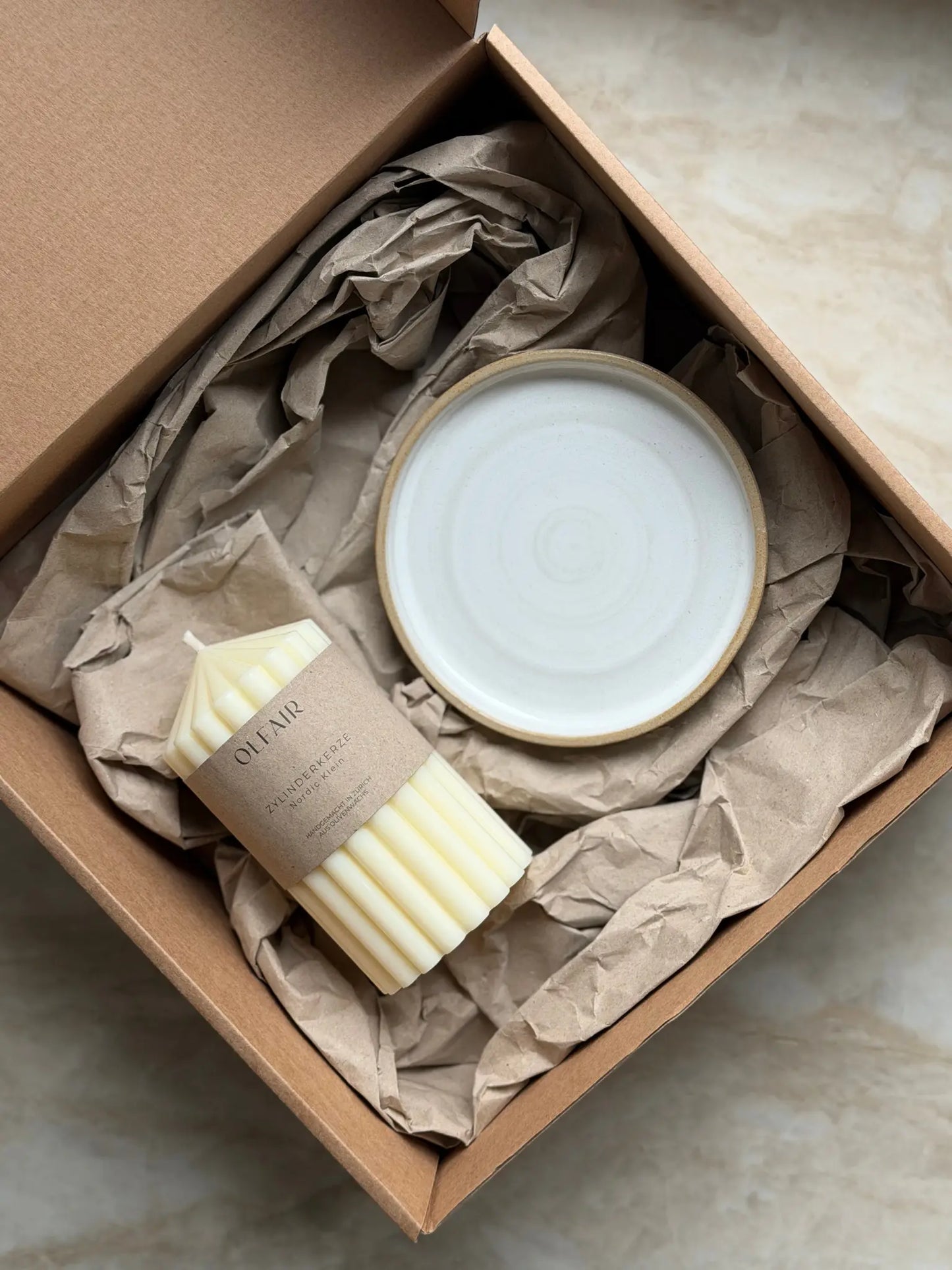 Geschenkset mit einer kleinen Zylinderkerze mit breiten Rillen und einem Kerzenteller aus Keramik in einer Kartonbox.