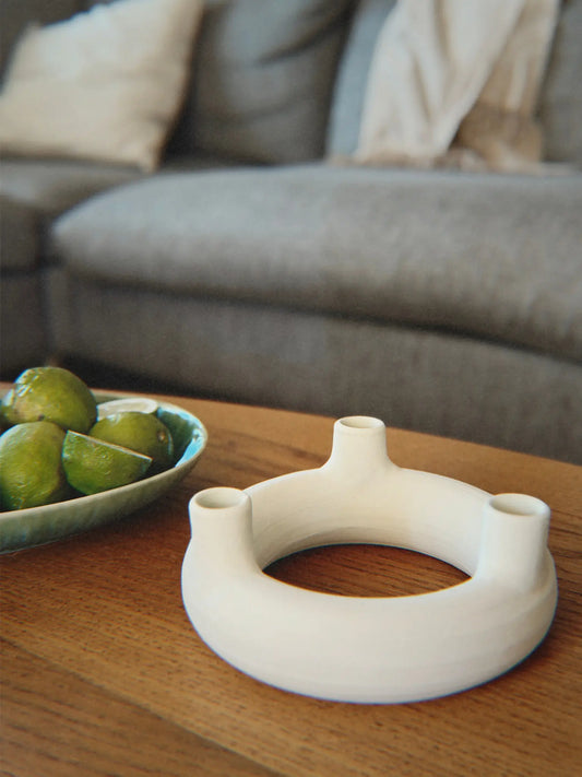 Runder Kerzenhalter mit Löchern für drei Kerzen aus unglasierter Keramik in off-white auf einem Couchtisch.