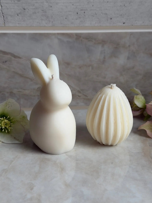 Osterhasen Kerze mit beiden Ohren und Ostereier Kerze mit Streifen vor einem Marmor Hintergrund.