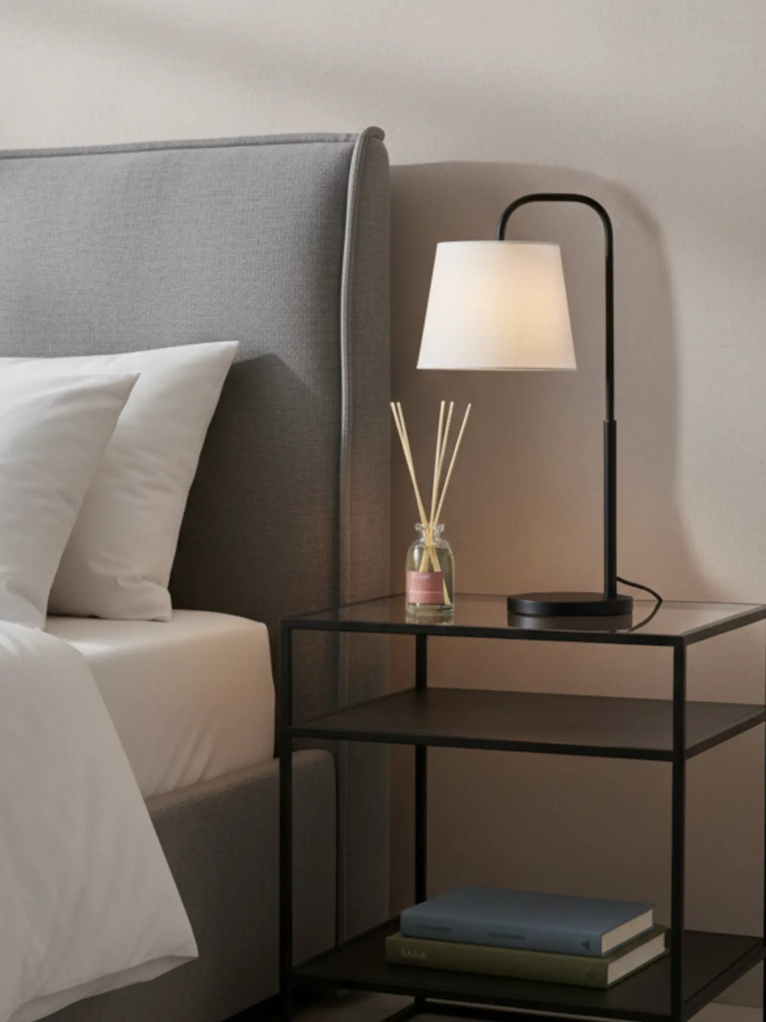 Ein Schlafzimmer mit einem grauen Bett mit mit weisser Bettwäsche. Daneben steht ein schwarzer Nachttisch aus Metall. Auf der unteresten Etagen liegen zwei Bücher. Auf der obersten steht eine Nachttischlampe und ein Raumduft Flower Meadow.