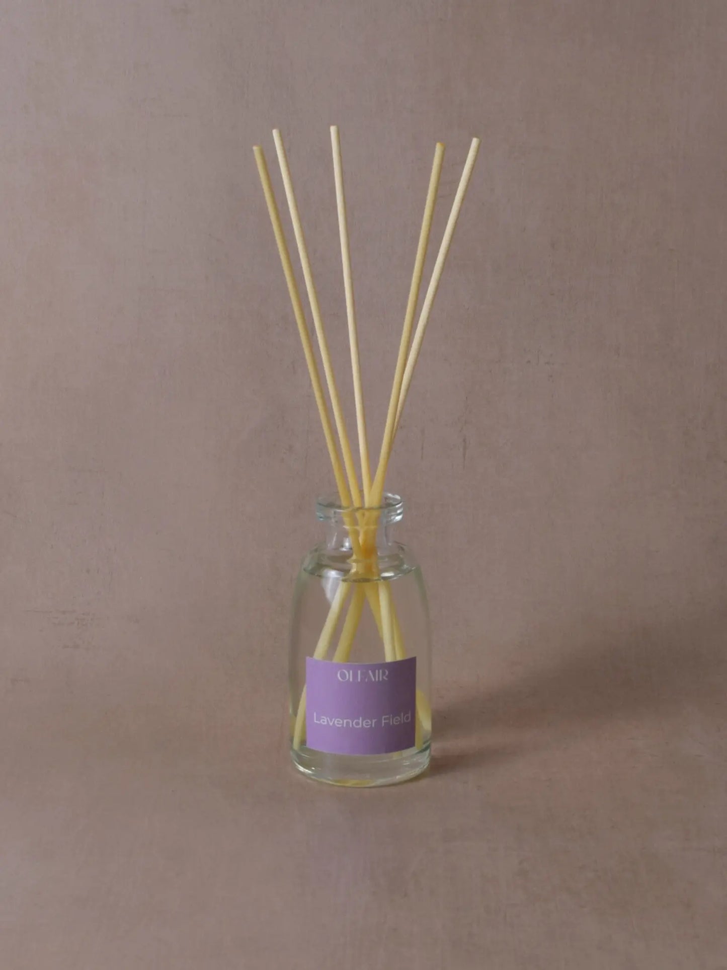 Kleiner Raumduft mit Stäbchen und einem violetten Etikett vor einem beigen Hintergrund.