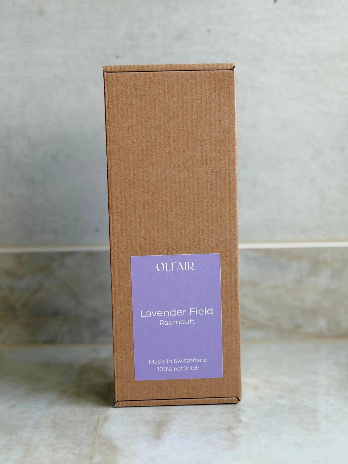 Kartonbox für Raumduft mit violettem Etikett vor einem Marmor Hintergrund.