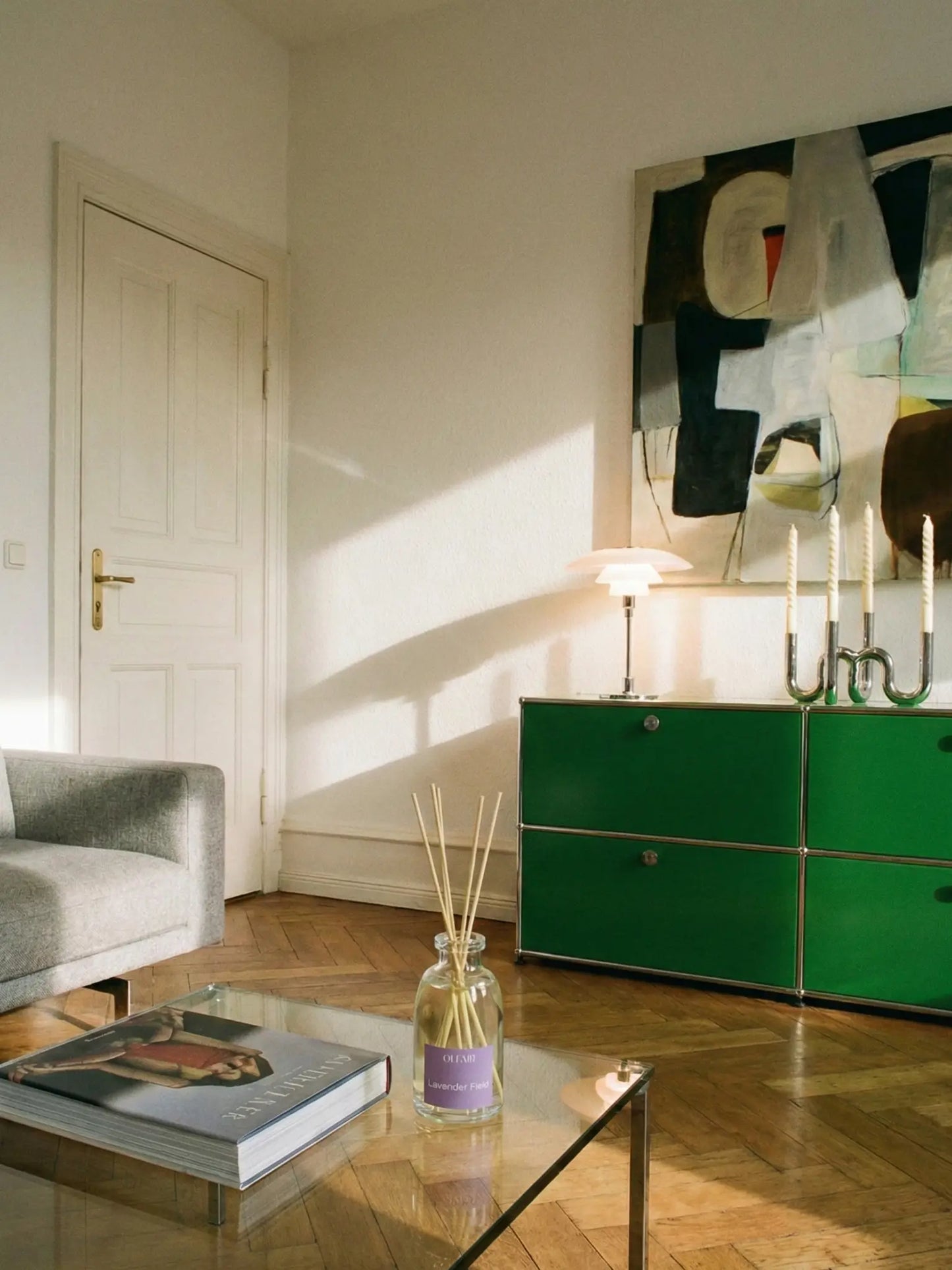 Wohnzimmer in einer Albauwohnung. Von einer Seite scheint Sonnenlicht ins Zimmer. An der Wand steht ein grünes Sideboard. Darauf steht eine kleine Lampe und ein Kerzenständer aus Metall mit vier weissen Kerzen. Darüber hängt ein abstraktes BIld. In der Mitte steht ein Glas Couchtisch mit einem Buch und einem Raumduft darauf.