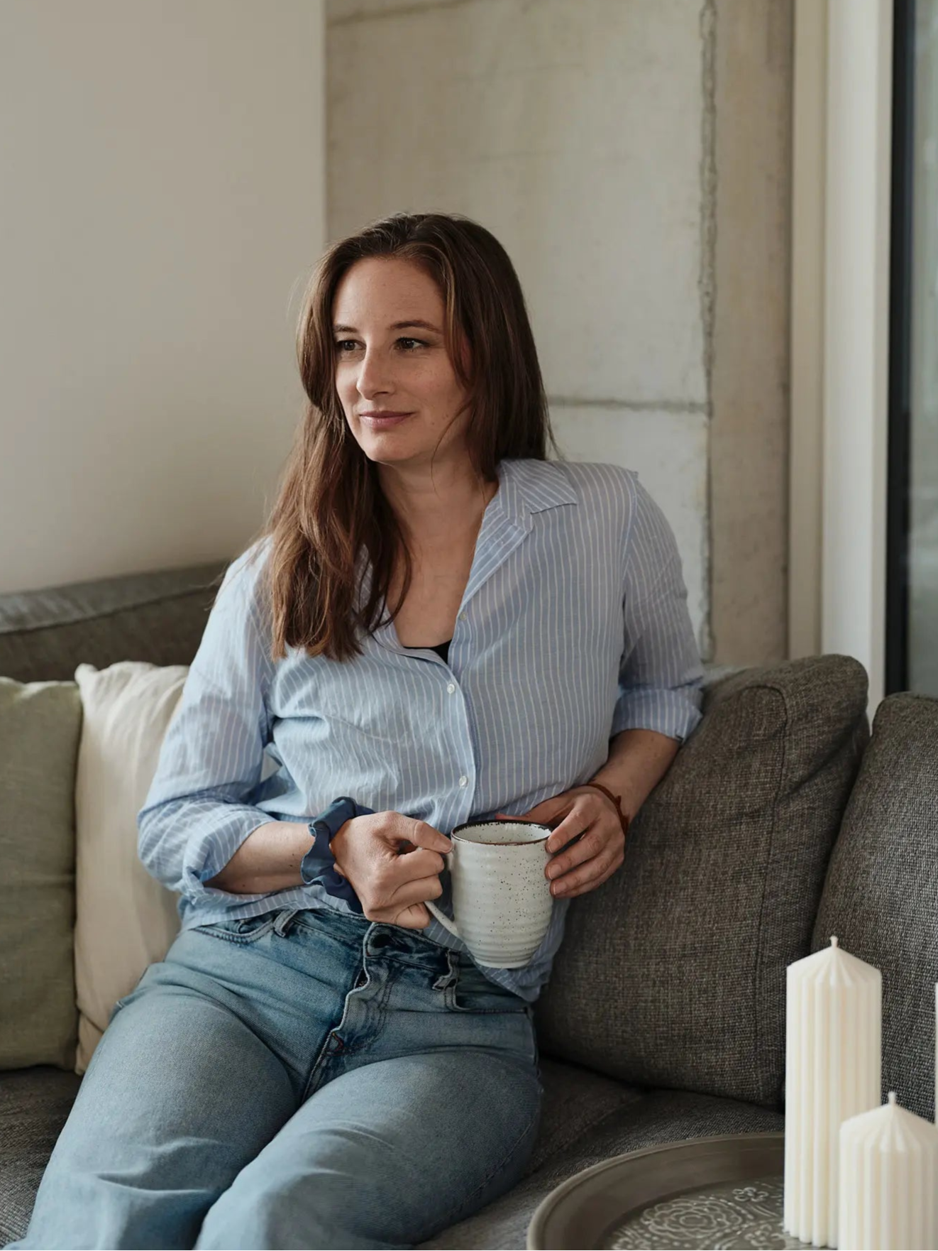 Jessica Niedermann, die Gründerin und Inhaberin von Olfair sitzend auf einem grauen Sofa mit Tee in der Hand und Zylinderkerzen als Dekoration