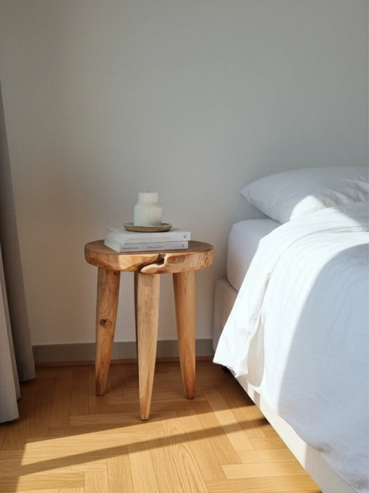 Ein Schlafzimmer mit einem weissen Bett und weisser Bettwäsche. Daneben steht ein kleiner Hocker aus Holz mit Büchern und einer Kerze mit feinen Rillen. Leichtes Sonnenlicht fällt ins Zimmer.