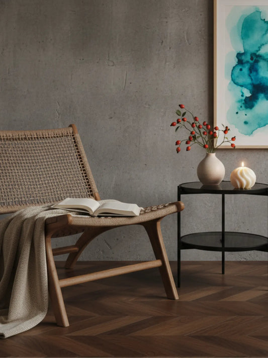 Ein Lounge Stuhl aus Holz in einem Wohnzimmer. Darauf liegt eine Wolldecke und ein aufgeschlossenes Buch. Neben dem Buch steht ein schwarzer Glastisch. Darauf steht eine weisse, runde Kerze mit Wellen und eine Vase mit Hagebuttenzweigen. An der Wand hängt ein Aquarellbild in türkis. Das Licht ist gemütlich.