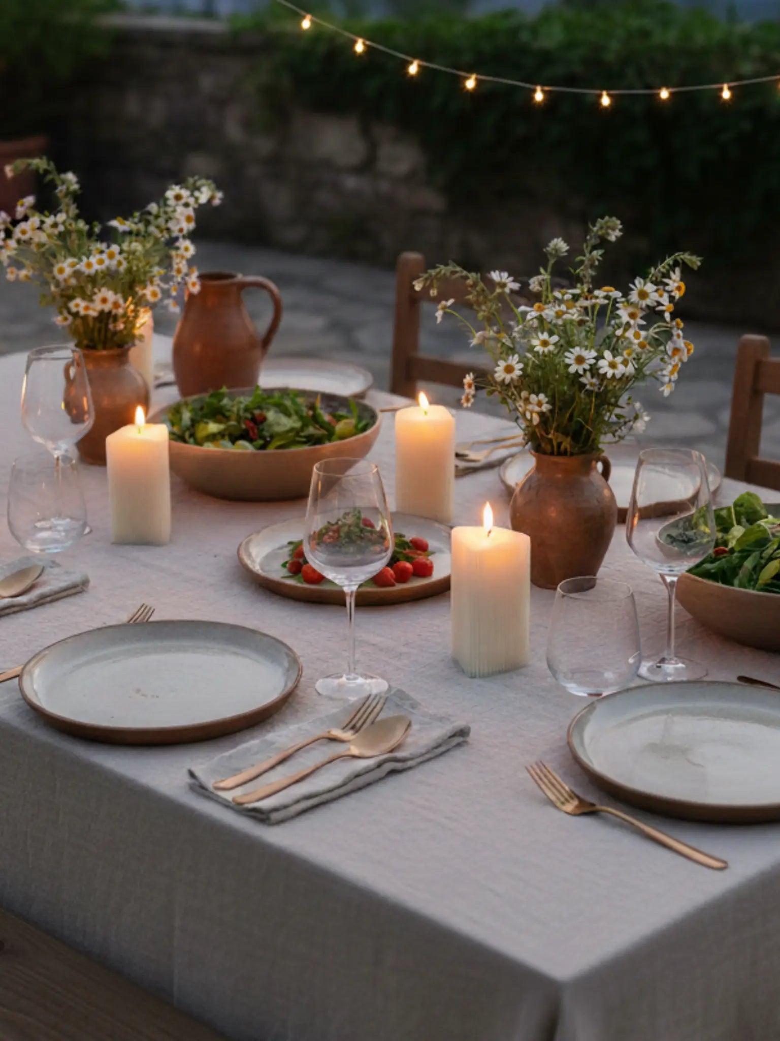 Kleine, dreieckige Kerzen mit feinen Rillen auf einem gedeckten Tisch mit Salat und Blumen. Der Tisch steht auf einer Terrasse. Es ist bereits am Eindunkeln.