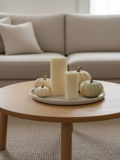 Grosse und kleine, dreiecke Kerzen auf einem weissen Kerzenteller auf einem hellen Holztisch. Daneben stehen einige Kürbise. Im Hintergrund ist ein helles Sofa zu sehen.