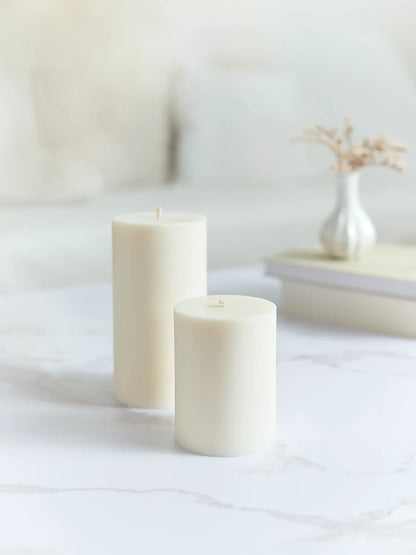 Eine grosse und eine kleine glatte Zylinderkerze in cremeweiss. Beide stehen auf einem Marmorcouchtisch und im Hintergrund hat es Bücher und eine Vase mit Trockenblumen.