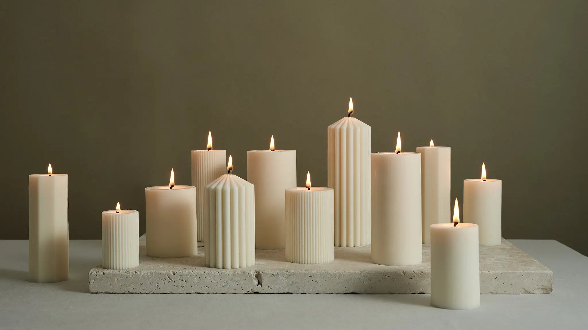 Kollektion aus elfenbeinfarbenen Stumpenkerzen in verschiedenen Höhen und geriffelter Struktur, brennend auf einer Natursteinscheibe vor warmem, neutralem Hintergrund arrangiert.